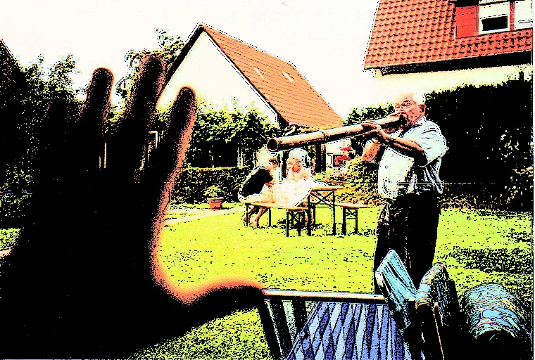 Stark kontrastreiches Foto eines Gartens: rechts steht ein älterer Mann in hellem Hemd und Weste und bläst in ein langes, waagerecht gehaltenes Rohr. Dahinter Rasen, eine Bierzeltgarnitur mit zwei sitzenden Personen, Hecken, ein weißes Haus mit spitzem Giebel und rechts ein Dach mit roten Ziegeln. Links ragt eine große, unscharfe dunkle Hand mit gespreizten Fingern ins Bild.