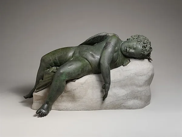 Bronzefigur eines schlafenden Kindes mit Flügeln, ausgestreckt auf einem hellen Steinblock liegend, den Kopf auf den angewinkelten Arm gelegt, ein Bein hängt über die Kante; die Oberfläche ist grün angelaufen.
