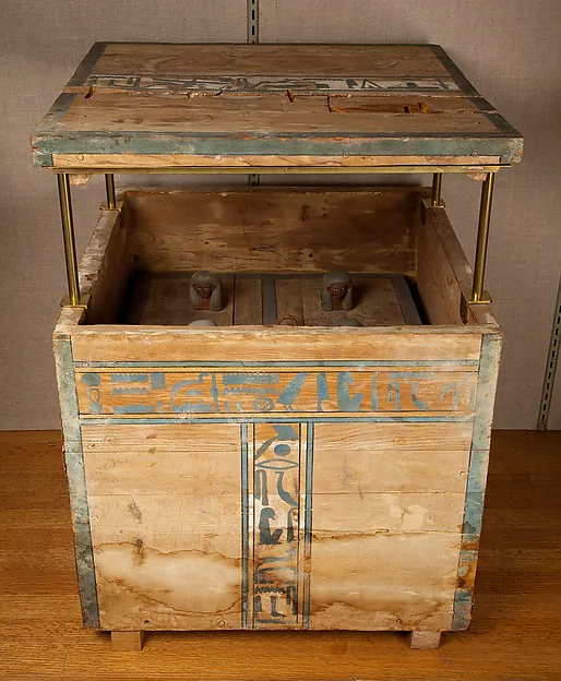 Bemalte Holzkiste mit Hieroglyphen in Blau und Ocker; der abgenommene Deckel schwebt an Metallstangen darüber, im Inneren stehen kleine Gefäße mit menschen- und tierköpfigen Verschlüssen.