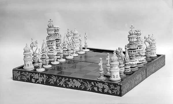 Schwarzweißfotografie: zwei Gruppen hoher, reich geschnitzter heller Spielfiguren stehen an den gegenüberliegenden Rändern eines dunklen Klappbretts mit Blumenintarsien; das karierte Feld in der Mitte ist leer.