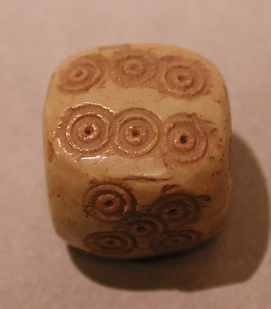 Ein einzelner, stark abgerundeter Würfel aus hellem Knochen; die Augen sind als eingeritzte konzentrische Kreise ausgeführt, drei Flächen sind sichtbar.
