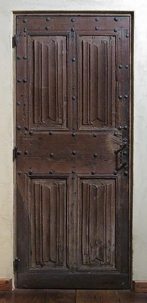 Schmale dunkle Holztür mit vier Füllungen und Reihen eiserner Nägel, eingelassen in eine helle Wand über einem Dielenboden.