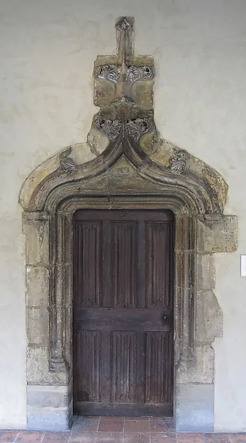 Steinernes Spitzbogenportal mit verwittertem Maßwerk in einer verputzten Wand, darin eine geschlossene dunkle Holztür mit schmalen Kassetten.