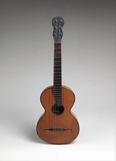 Eine kleine Gitarre mit hellem Holzkorpus, schmalem Hals und dunklem Griffbrett, aufrecht vor grauem Hintergrund; am Kopf sitzt eine metallene Stimmmechanik.