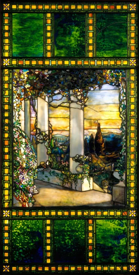 Bleiverglastes Fenster aus Buntglas: ein breiter Rahmen aus Reihen kleiner bernsteinfarbener und grüner Glasquadrate umfasst in der Mitte den Blick durch eine Pergola mit hellen Säulen, die von blühenden Ranken überwachsen sind; dahinter eine Brüstung, eine dunkle Zypresse, Hügel und ein gelb-blau geschichteter Himmel; über und unter dem Bild große Felder aus wolkig grünem Glas.