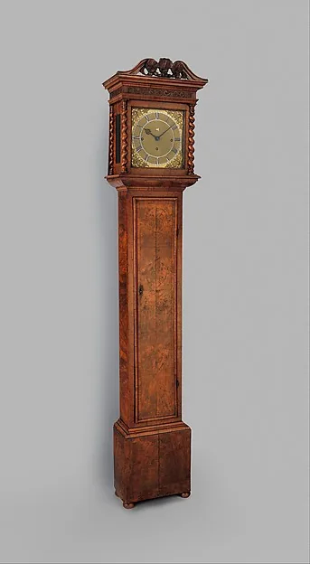 Hohe Standuhr aus geflammtem Nussbaumholz mit geschnitztem Aufsatz; oben ein quadratisches Messingzifferblatt, darunter eine schlichte lange Gehäusetür.