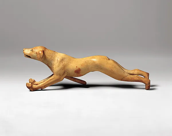 Eine langgestreckte Hundefigur aus hellem Elfenbein mit geöffnetem Maul und weit nach vorn und hinten gestreckten Läufen, freigestellt vor grauem Grund.