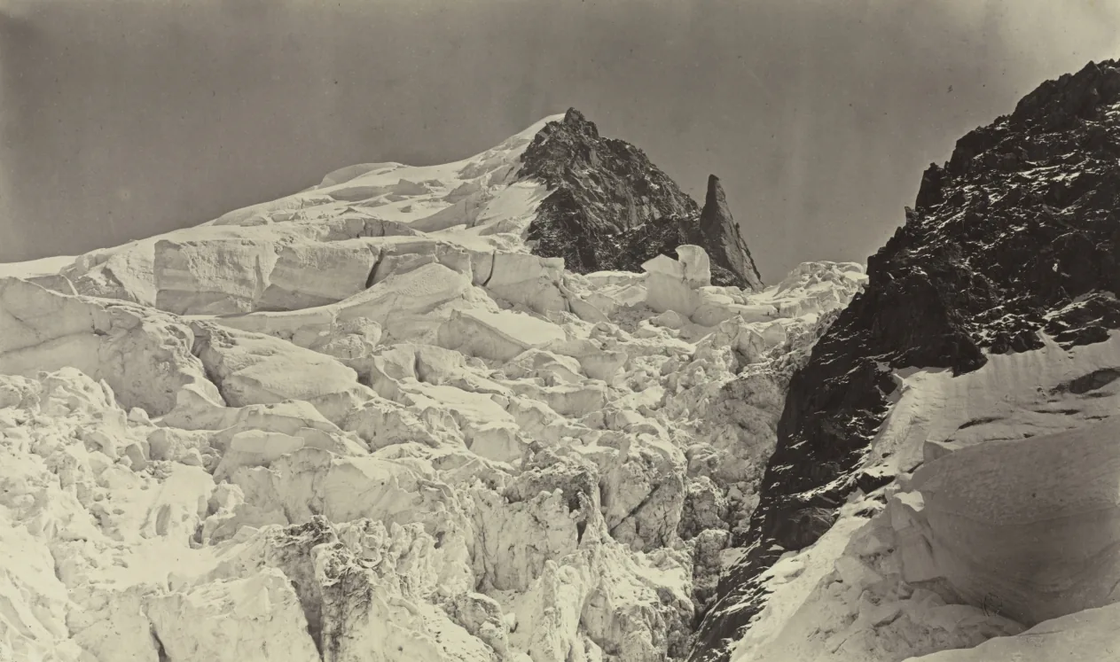 Schwarzweißfotografie: ein zerklüftetes Gletscherfeld aus gestuften Eisblöcken füllt den Vordergrund, dahinter ragt ein dunkler, kantiger Felsgipfel aus dem Schnee, rechts fällt eine schwarze Felswand ab, der Himmel darüber ist hell und leer.