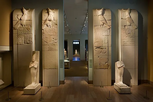 Museumssaal: vier hohe Kalksteinplatten mit Hieroglyphen und je einem Falken am oberen Rand stehen aufgereiht, dazwischen zwei kleine Figurenskulpturen und ein beleuchteter Durchgang in weitere Räume.