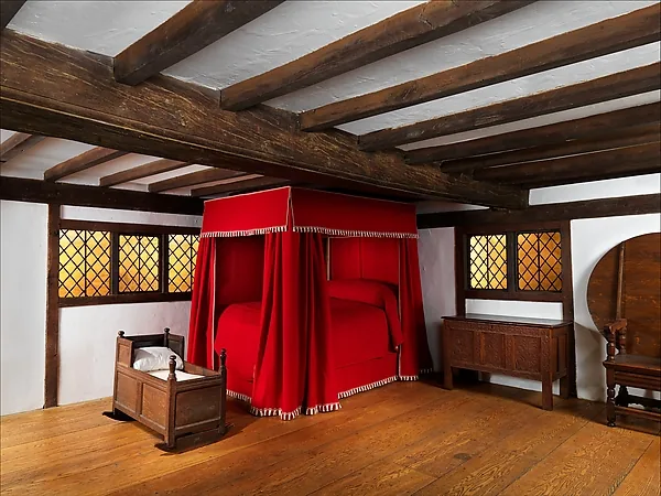 Zimmer mit dunklen Deckenbalken und breiten Holzdielen: in der Mitte ein Bett mit rotem Baldachin und geschlossenen Vorhängen, links eine hölzerne Wiege, rechts unter einem Butzenfenster eine niedrige Truhe und ein Stuhl mit runder Lehne.