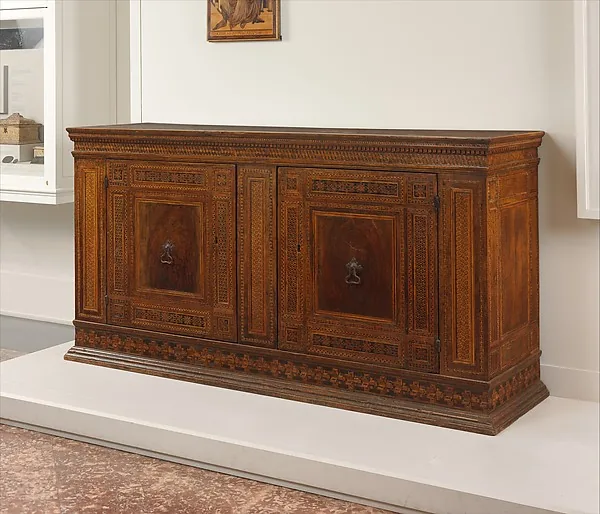 Langer niedriger Holzschrank mit zwei Türen und feiner geometrischer Einlegearbeit, aufgestellt auf einem hellen Sockel in einem Ausstellungsraum.