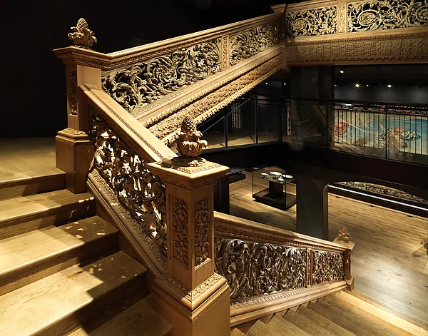 Fotografie einer hölzernen Treppe: das Geländer besteht aus dicht durchbrochenem, geschnitztem Blattwerk mit Zapfen auf den Pfosten; die Stufen laufen nach links aus dem Bild, ringsum ein dunkler Ausstellungsraum.