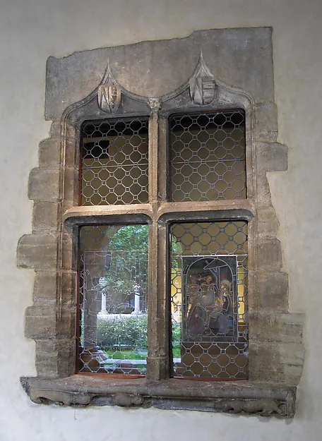 Fotografie: ein steinernes Spitzbogenfenster sitzt in einer hell verputzten Wand; die vier Felder sind mit bleigefassten Wabenscheiben verglast, in einem unteren Feld ein farbiges Glasbild mit Figuren, durch die übrigen Scheiben sieht man Grün und einen Hof.